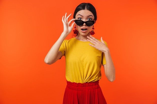 cheerful hispanic woman 20s wearing sunglasses smiling at camera isolated over red wall