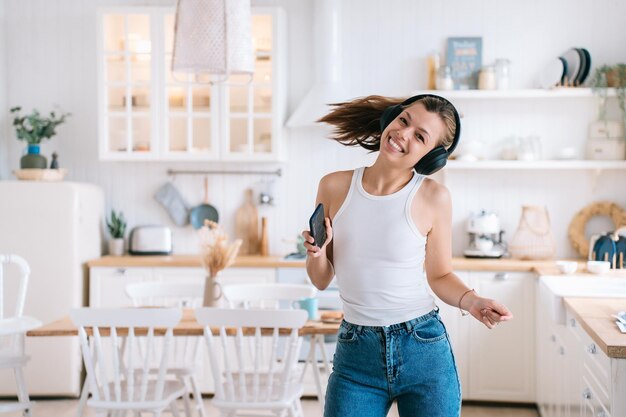 Cheerful hispanic young woman in casual using headphones and phone dancing at kitchen looks at camera toothy smiles happy by new home Beautiful spanish housewife having fun at home Domestic leisure