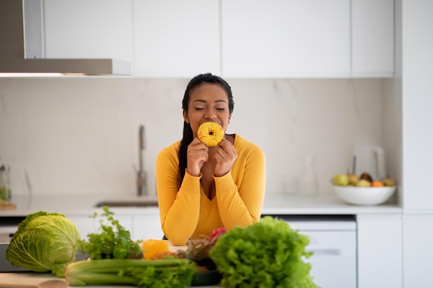 Cheerful hungry millennial african american lady in casual hold donut enjoy aroma at table with organic vegetables