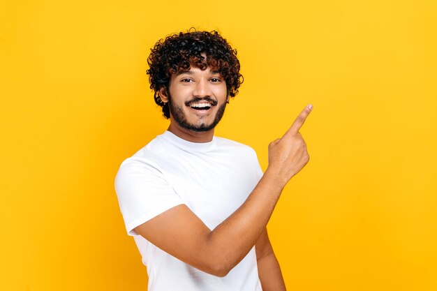 白い基本Tシャツを着た陽気なインド人またはアラビア人男性は見た目で指を横に指さしプレゼンテーションのためのモックアップスペースで孤立したオレンジ色の背景に立って笑顔でカメラを見ています