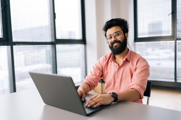 Cheerful Indian business man in eyeglasses sitting at desk with laptop smiling looking at camera in light coworking office by window