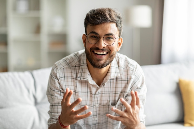 Photo cheerful indian guy talking looking at camera and gesturing communicating online sitting on couch at