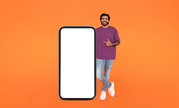 Cheerful indian man hipster pointing at big smartphone mockup