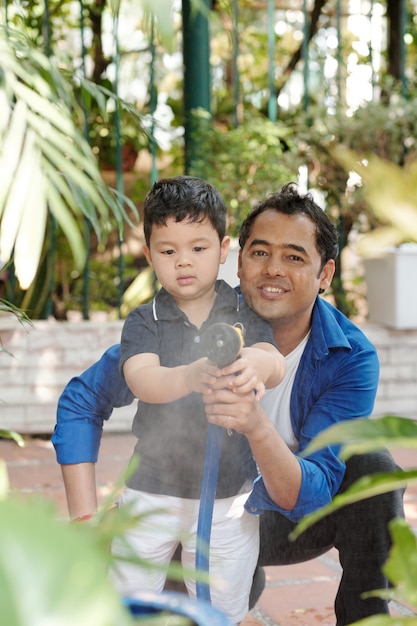 Cheerful Indian man and his little son standing in backyard and spraying water from garden hose