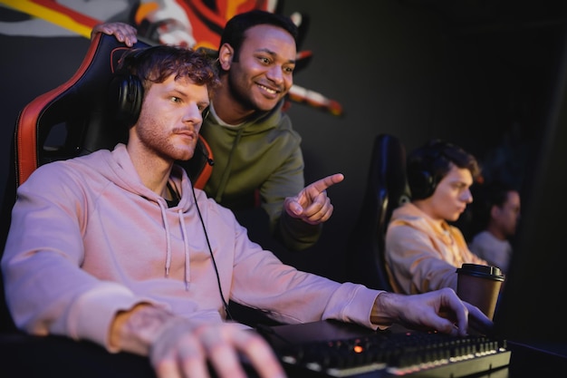 cheerful indian man pointing at computer while friend playing video game in cyber club