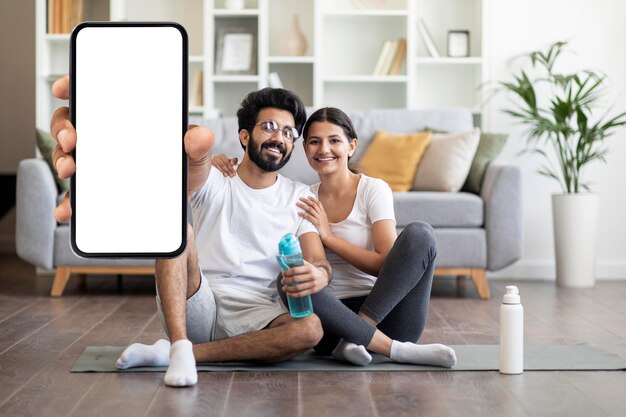 Cheerful indian spouses showing blank smartphone while training together at home