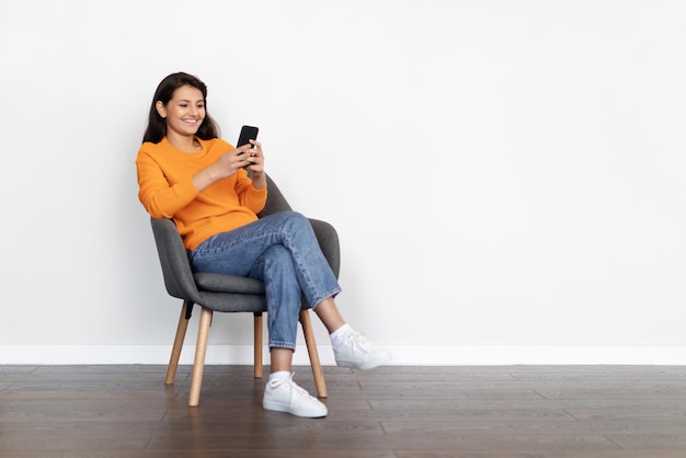 Cheerful indian woman sitting in armchair using cell phone