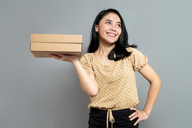 Cheerful Indonesian woman holding cardboard box with one hand on waist