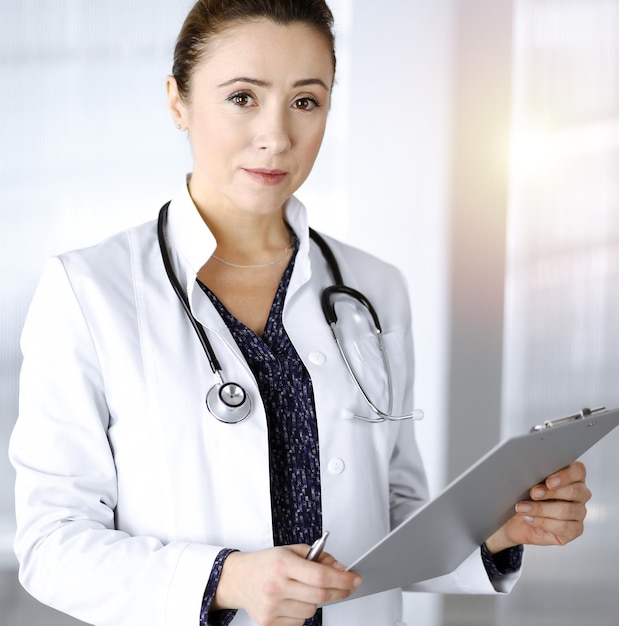 Cheerful intelligent woman-doctor is holding a clipboard in her hands, while standing in a sunny clinic office. Portrait of friendly physician woman with a stethoscope. Perfect medical service in a ho