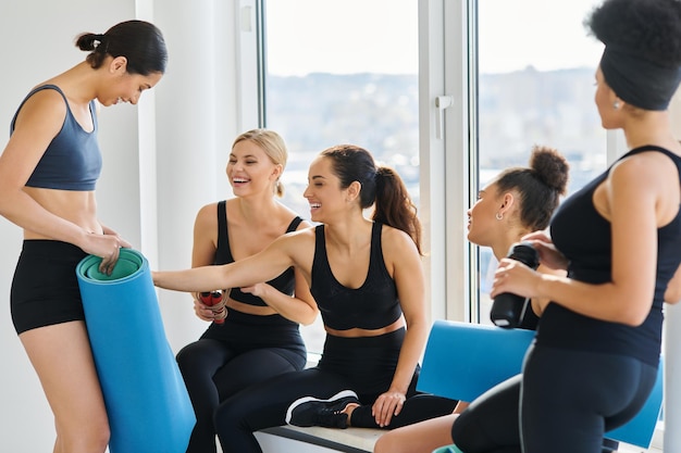 Foto giovani donne interrazziali allegre di vent'anni che chiacchierano sedute accanto alla finestra nello studio di pilates