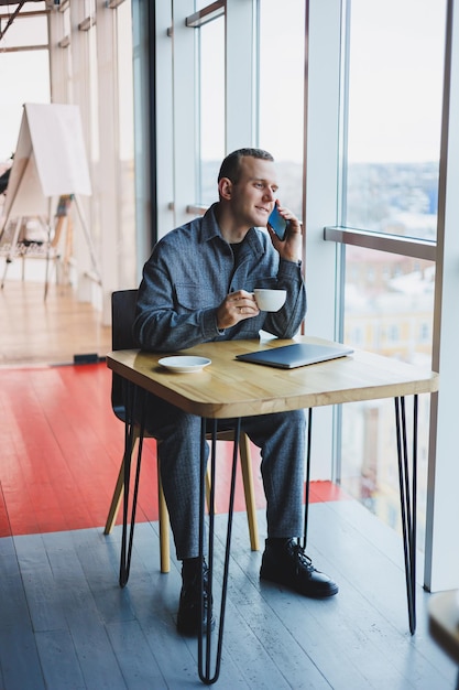 Cheerful IT professional with laptop talking on smartphone to discuss web programming happy businessman in cafe drinking coffee freelancer