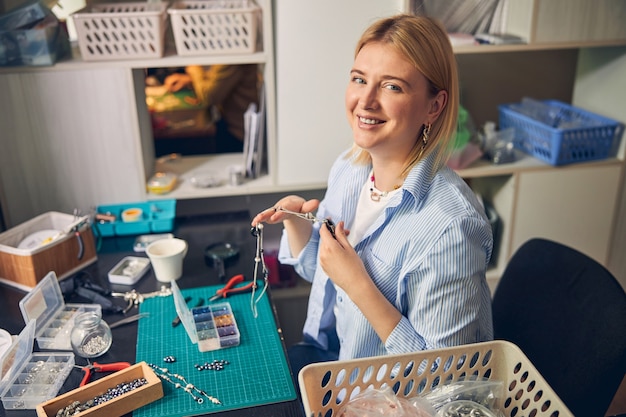 Cheerful jewel employee holding fashionable necklace while glancing at camera