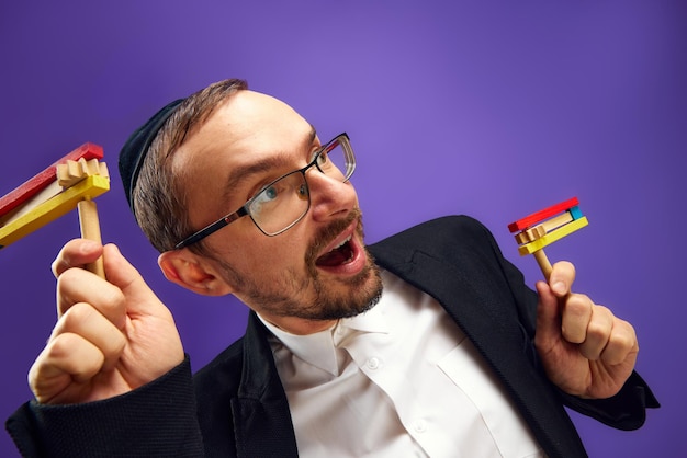 Cheerful jewish man in yarmulke and glasses holding noisemaker against purple studio background