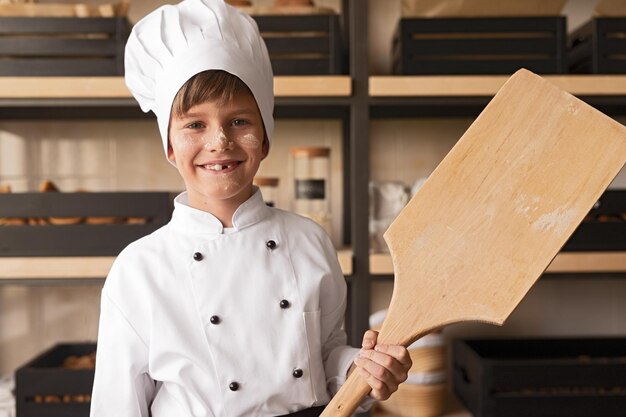 Cheerful kid in cook uniform with shovel in bakery