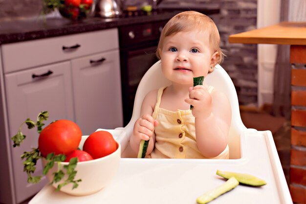 Cheerful kid eating vegetables. Portrait of a happy girl in a highchair in the kitchen