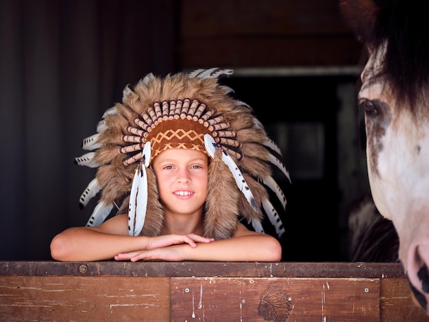 Cheerful kid wearing authentic Native American headwear leaning on barrier of stable near crop horse and looking at camera