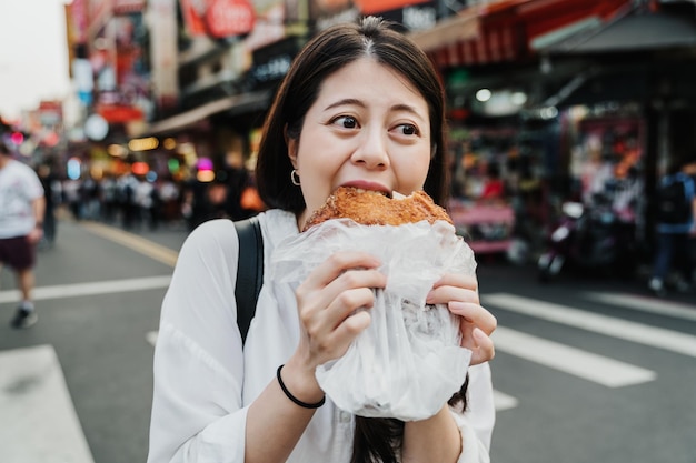 L'allegro viaggiatore con zaino e sacco a pelo della signora coreana viaggia a taiwan e prova il delizioso cibo di strada locale. ragazza che tiene un filetto di pollo amico all'interno del sacchetto di plastica e mangia con un sorriso. la bella donna ama la cucina