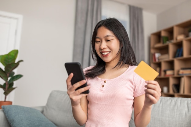 Cheerful korean lady with mobile phone and credit card purchasing goods or services on web sitting