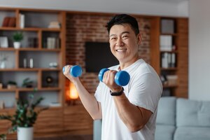 cheerful korean mature man doing strength exercises for arms lifting dumbbells in living room smiling at camera