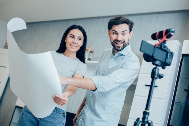 Cheerful lady holding large sheet of drawing paper while standing near the smiling man and being recorded on a camera