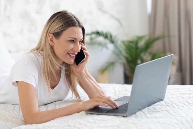 Cheerful lady independent contractor using notebook and smartphone in bed