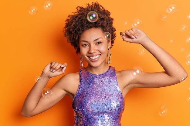 Cheerful lady with curly hairstyle dances at party shakes arms smiles happily has good mood dressed in glittering purple t shirt isolated over orange wall soap bubbles around enjoys good day