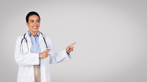 Cheerful latin female doctor in workwear coat pointing fingers aside at copy space for medical