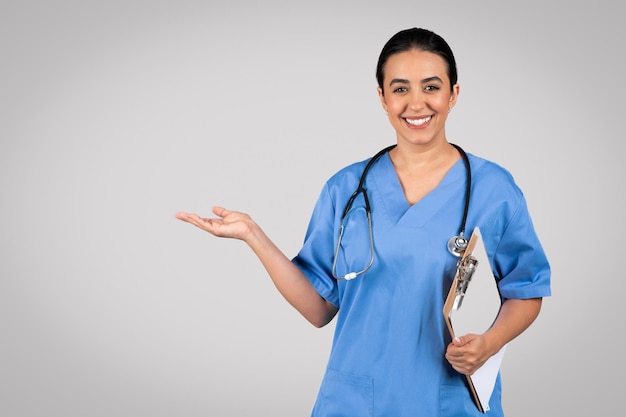 Cheerful latin woman doctor in blue workwear holding medical chart pointing at empty space showing