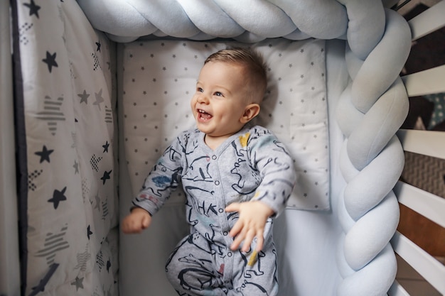 Cheerful laughing blond little baby boy with beautiful blond eyes rolling in his crib. Morning time.