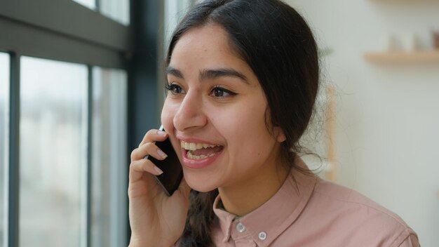Веселая смеющаяся индийская арабская девушка студентка Gen Z говорит по мобильному телефону в офисе небрежно