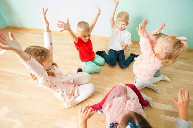 Bambini allegri e ridenti seduti su un pavimento in cerchio con le mani alzate. bambini in età prescolare felici che si divertono al centro diurno.