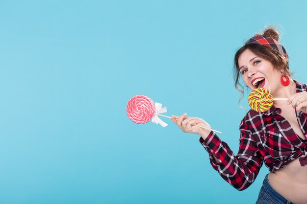 오른쪽을 보여주는 막대 사탕과 파란색 표면에 포즈 복고풍 옷에 쾌활한 웃음 예쁜 젊은 여자