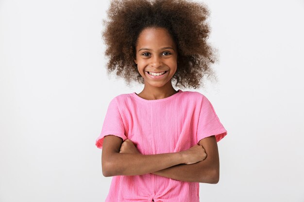 Cheerful little african girl wearing pink blouse standing isolated over white wall, arms folded