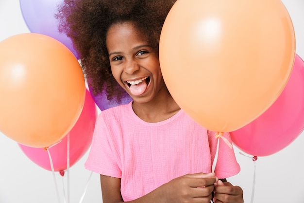 Cheerful little african girl wearing pink blouse standing isolated over white wall, having fin with air balloons, sticking her tongue out