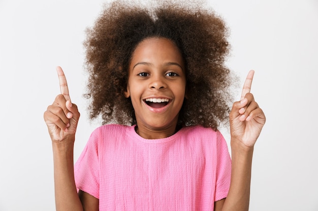 Cheerful little african girl wearing pink blouse standing isolated over white wall, pointing fingers at copy space