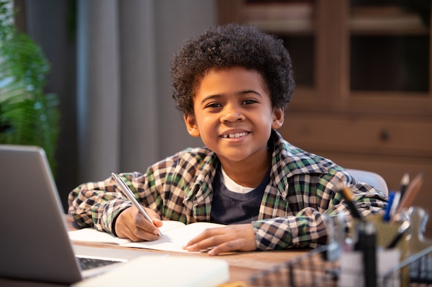 Cheerful little African schoolboy making notes in copybook and looking at you while sitting by table in front of laptop in home environment