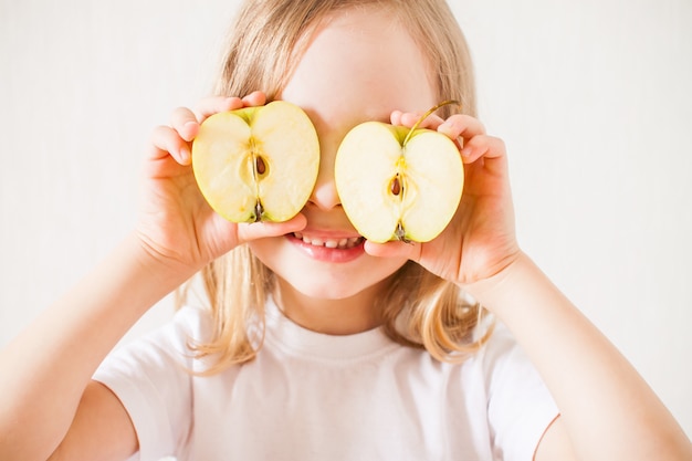 A cheerful little blonde girl smiling, having fun and looking through two halves of an apple in front of her eyes