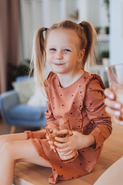 Cheerful little blonde Swedish girl with tails sitting on table holds a cookie glass of milk