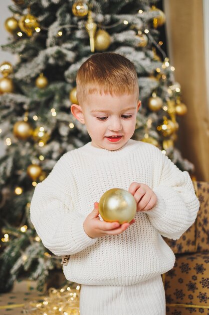 23歳の陽気な小さな男の子は ⁇ 花束に囲まれたクリスマスツリーの近くに立っています ⁇ クリスマスはクリスマス物語のコンセプトを提示します ⁇ 幼児のための休日 ⁇ クリスマスツリー