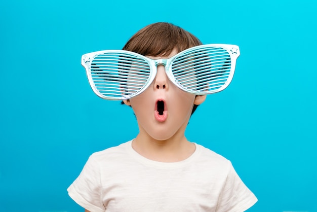 Cheerful little boy in big glasses express a surprised face isolated on blue wall