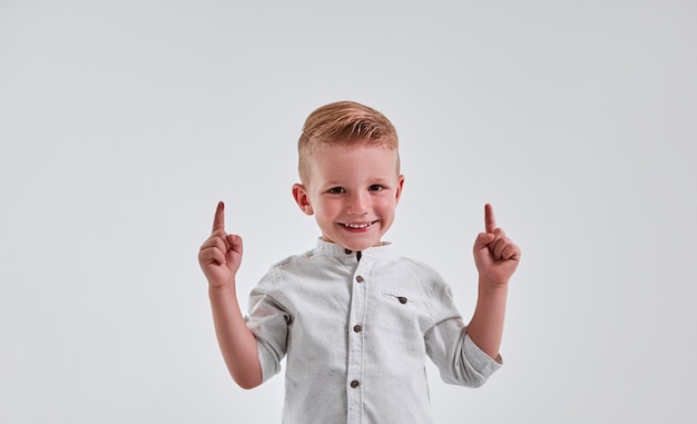 Cheerful little boy is pointing up with both hands over gray background and smiling.