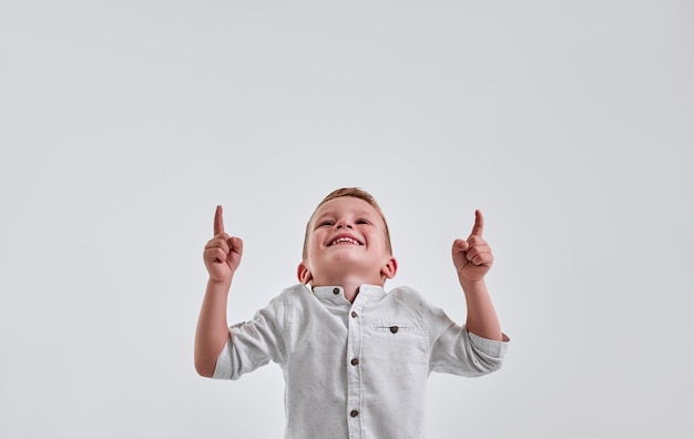 Фото Веселый маленький мальчик смотрит и указывает вверх обеими руками на сером фоне и улыбается.