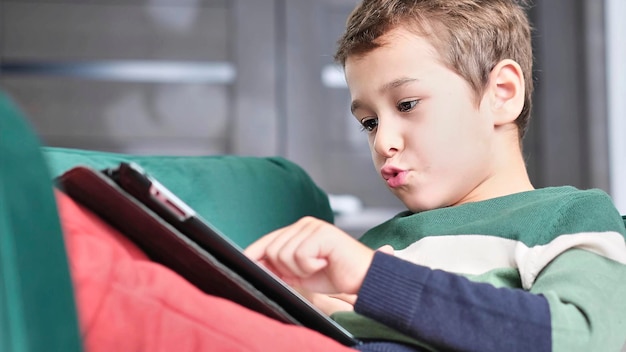 Cheerful little boy smiling while sitting on couch and using tablet at home Modern kid and education technology little boy looking at touch pad at home