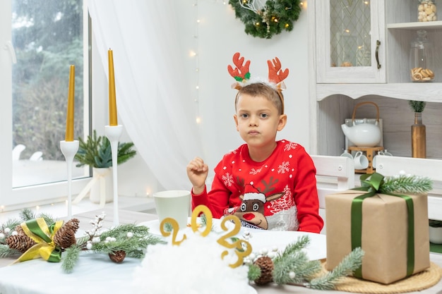 Веселый маленький мальчик в красном рождественском свитере с оленьими рогами на голове ест сухие дольки апельсина