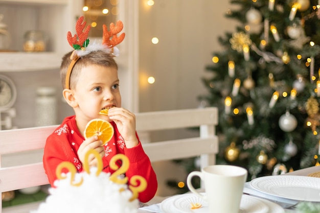 Cheerful little boy wearing warm red xmas sweater deer horns on head eat dry orange in kitchen