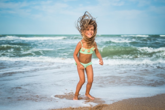 Cheerful little caucasian girl jumping and having fun on the sea waves on the sandy coast on a sunny hot summer day