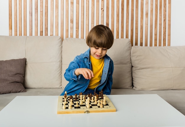 Cheerful little chess player in a denim shirt sits at a table and plays chess in a room