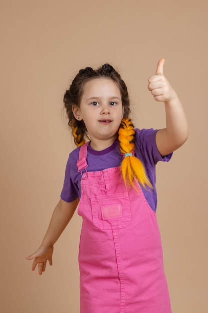 Cheerful little female showing like sign with hand having yellow kanekalon braids looking at camera wearing pink jumpsuit and purple tshirt on beige background