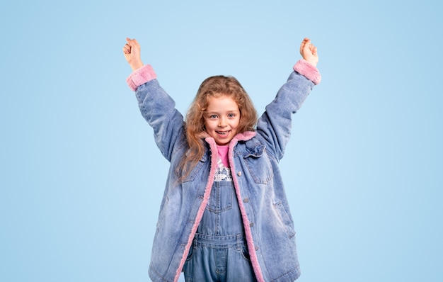 Cheerful little girl in blue warm denim jacket raising hands and looking at camera while standing against blue background