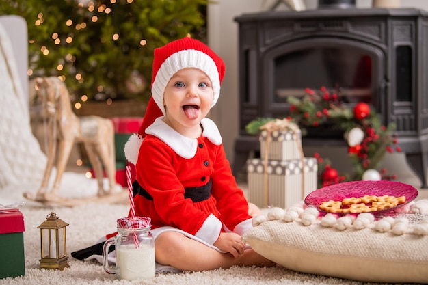 Cheerful little girl dressed as Santa Claus eats cookies and drinks milk in the living room on the background of the Christmas tree